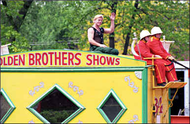 A strong man waves from a circus wagon.